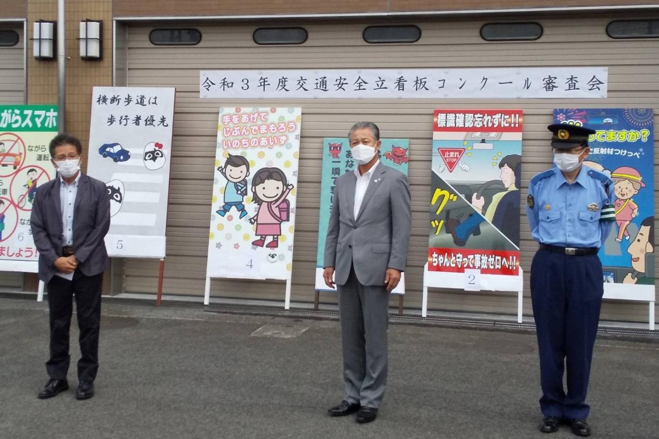 後方に6枚の交通安全立看板が設置され、3名の関係者が並んでいる写真