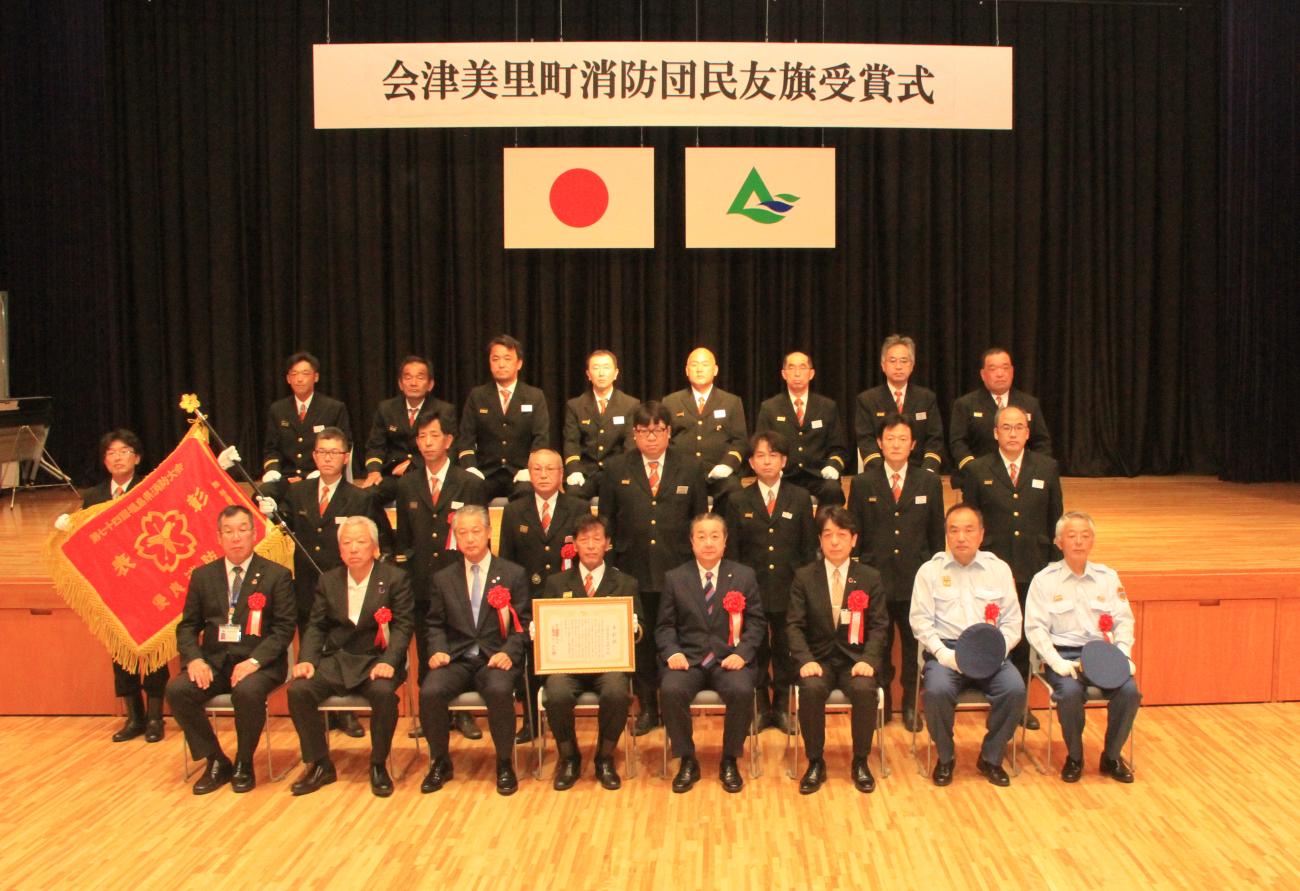 左脇に旗を持ち、制服を着た消防団の方々が横3列に並び、町長と一緒に記念撮影をしている写真