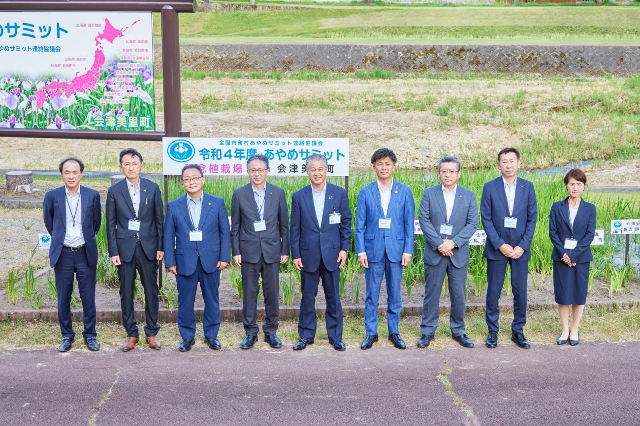 「あやめサミット」の看板を背景にスーツを着た9人の町長と関係者の方々が横一列に並び、記念撮影をしている写真