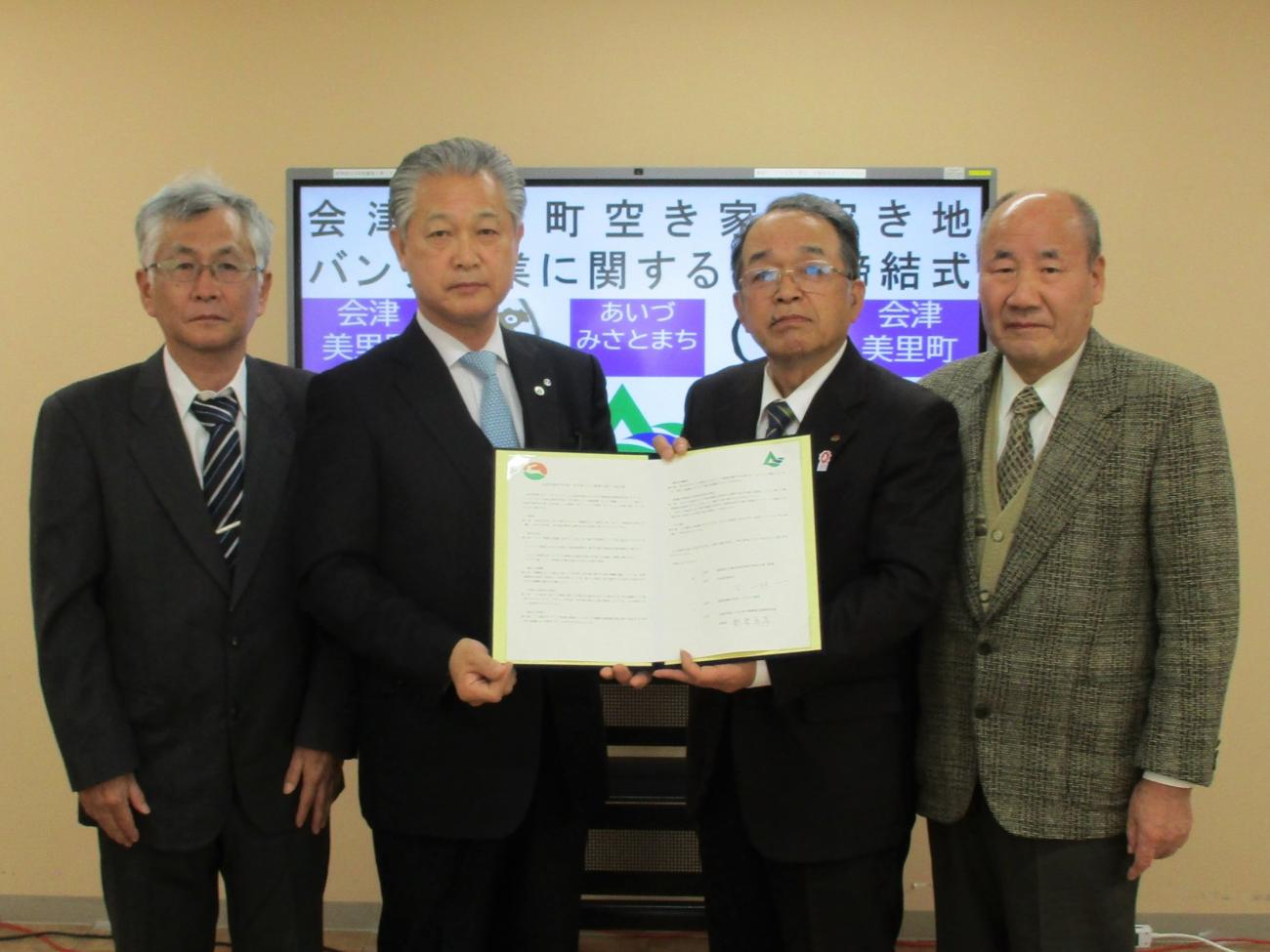 スーツ姿の男性4人が横一列に並び、中央の2人が一緒に協定書を持って記念撮影をしている写真