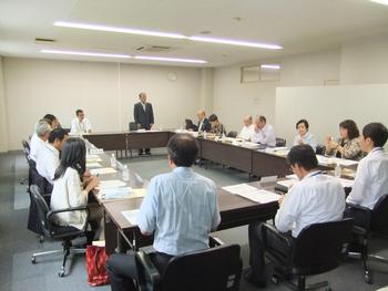 ロの字に設置された長机の席に多くの関係者が座り、前方の席の男性が立って話をしている様子の写真