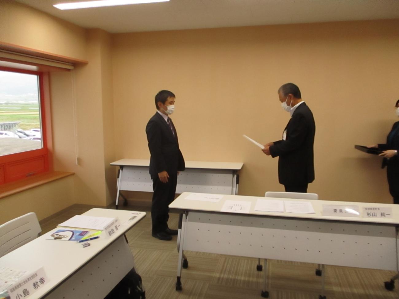 スーツ姿の男性が向かい合って立っており、右側の男性が委嘱状を手に持っている写真