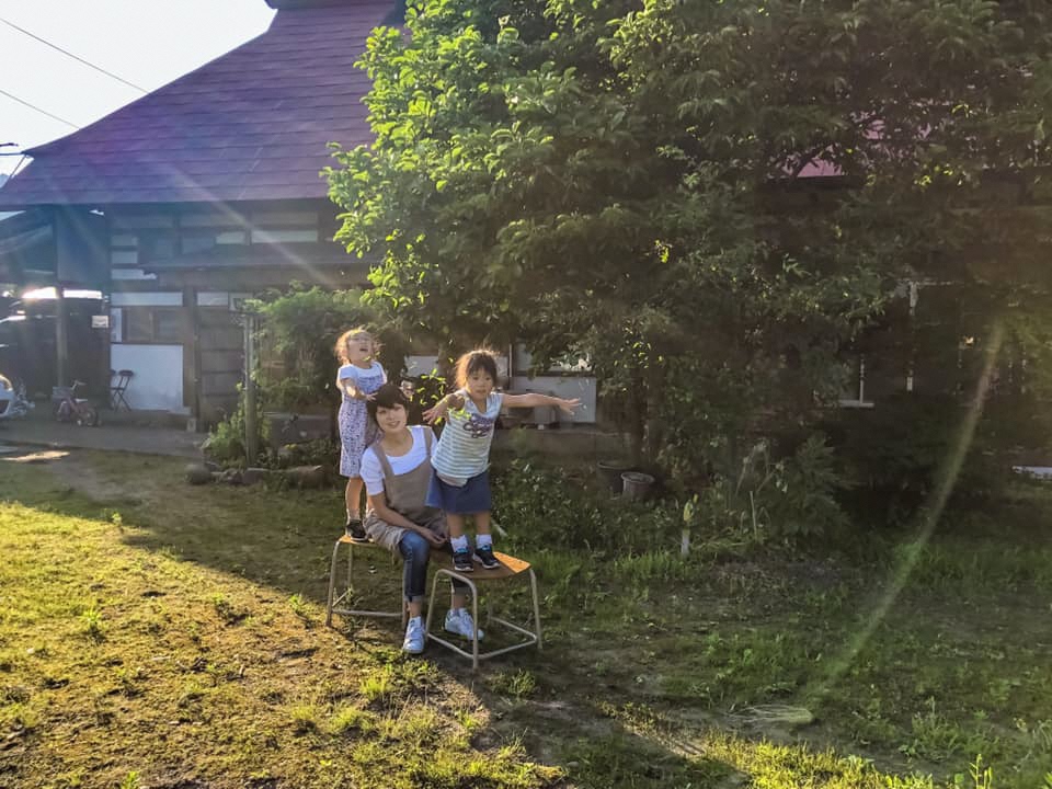 緑の庭に椅子を並べ、聡子(さとこ)さんの前後に娘さん2名が楽しそうに立っている写真