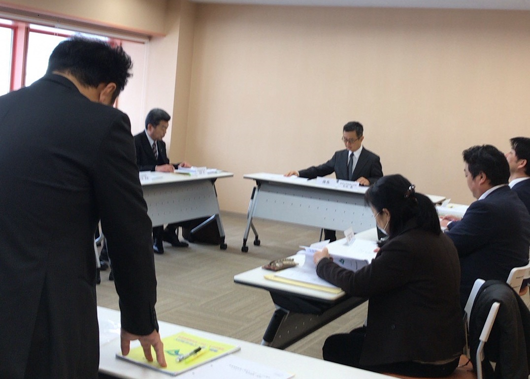 手前の男性が立って資料を見ている様子を後方から写した写真