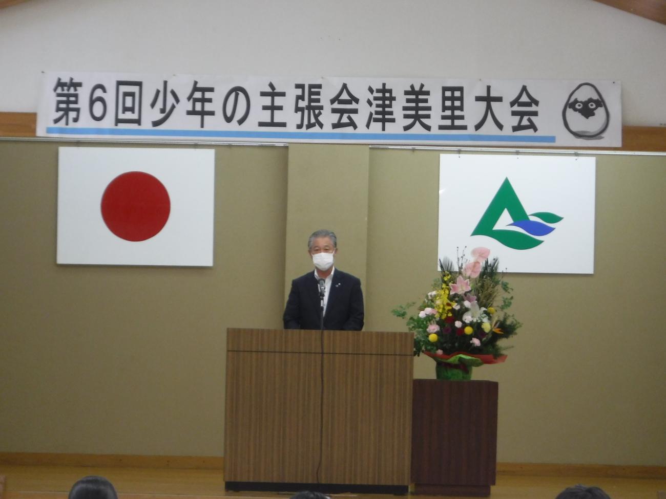 舞台に日本の国旗（左）と会津美里町の町章（右）が掲げてあり、演台に立ちマイクを通して話をしている町長の写真