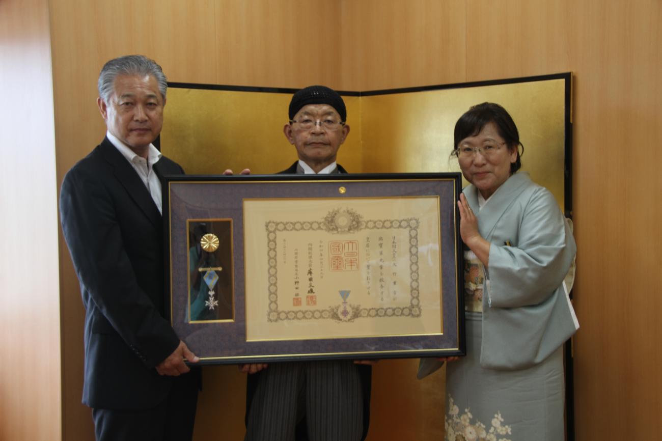 3人の男女が横1列に並び、町長（左）と大竹重吉さん（中央）と着物を着た女性が、額に入った賞状と勲章を一緒に持って記念撮影をしている写真