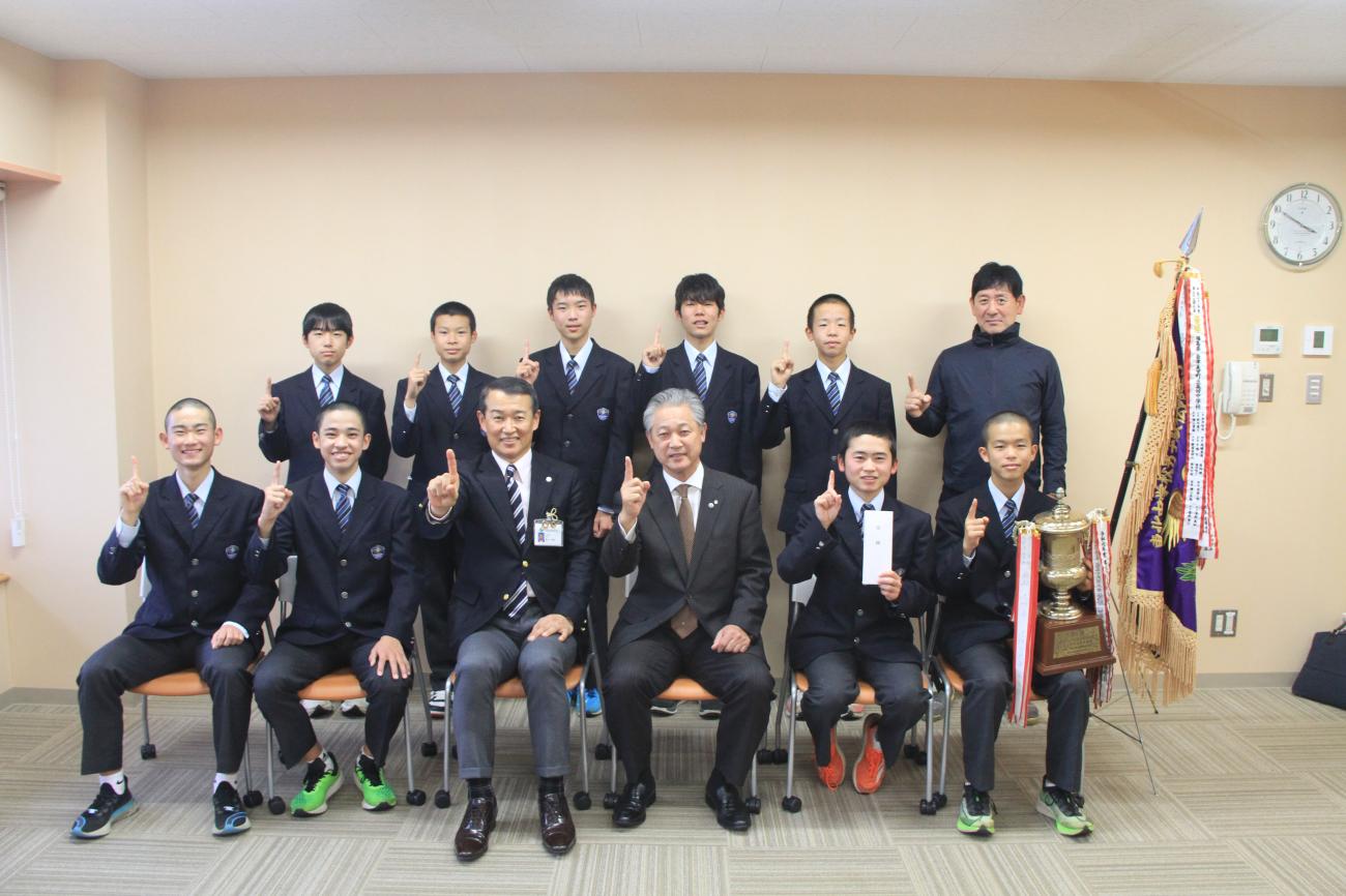 右側に優勝旗を置き、制服を着た高田中学校男子駅伝部の生徒と先生が町長と一緒に人差し指を上げるポーズをして記念撮影をしている写真