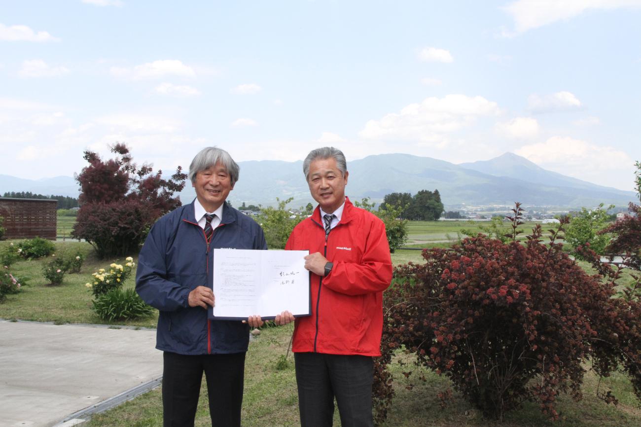 遠くに山並みが見える屋外で、株式会社モンベルの辰野勇様（左）と町長（右）が協定書を一緒に持ち、記念撮影をしている写真