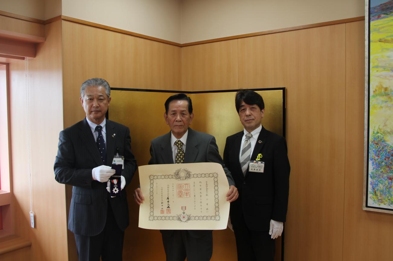 3人の男性が横1列に並び、町長（左）が勲章を持ち、歌川重雄さん（中央）が賞状を持って記念撮影をしている写真