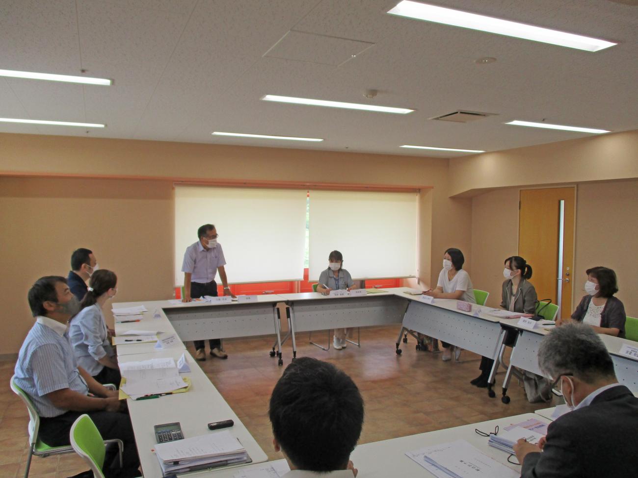 テーブルがロの字型に設置された会議室で、1人の男性が立って話をしており、自席に座っている関係者たちが男性の方を向いている写真