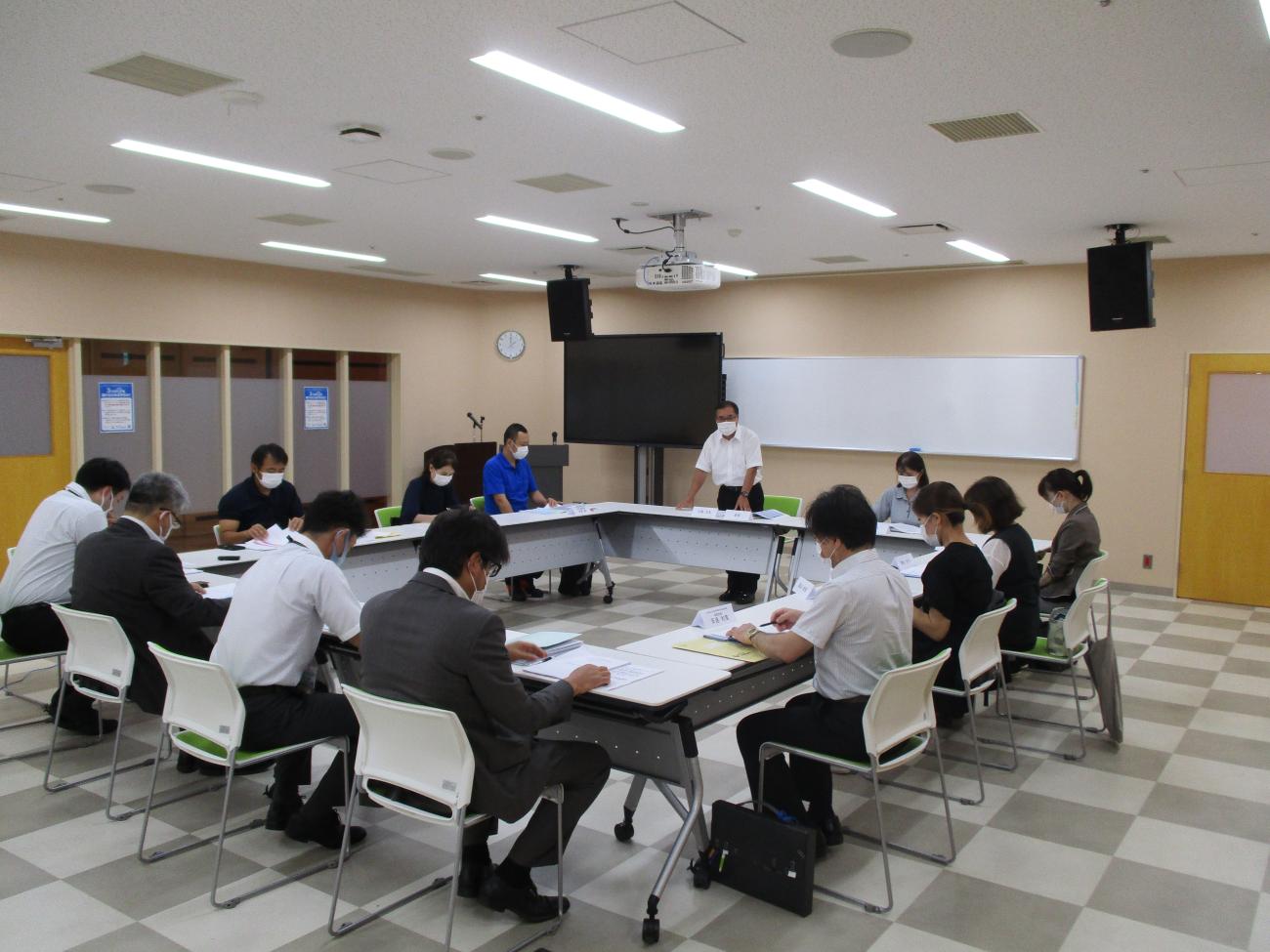 長テーブルがロの字型に設置された会議室で、部屋の正面側にいる男性が立っており、12人の関係者たちが自席に座って資料に目を通している写真