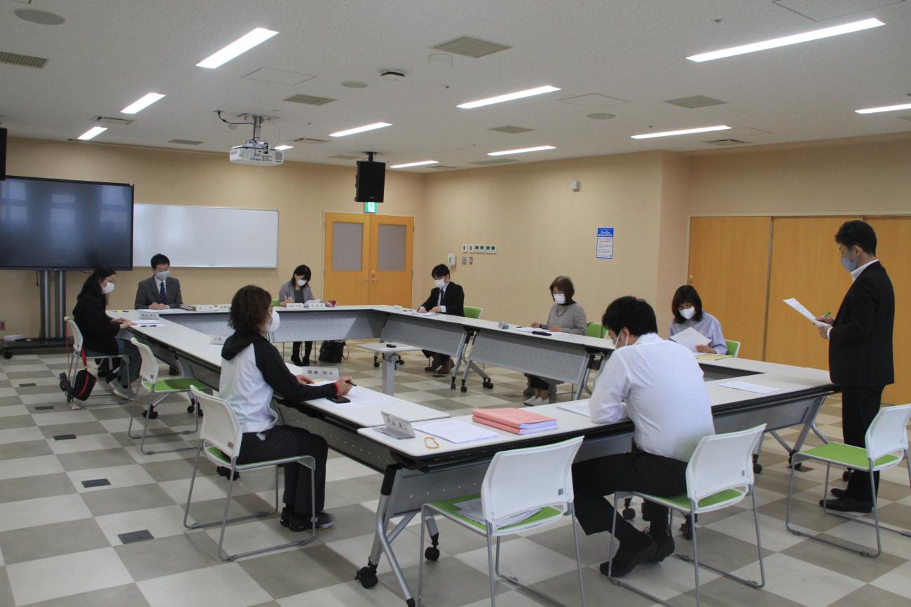 長テーブルがロの字型に設置された会議室で9人の関係者が会議しており、手前右側の男性が資料を持って立っている写真