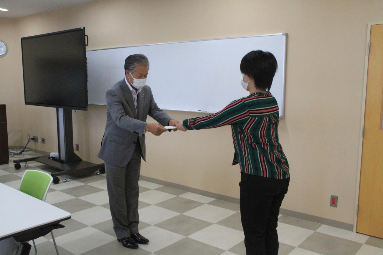 会議室の正面で、スーツ姿の男性が向かい合って立っている女性へ委嘱状を交付している写真