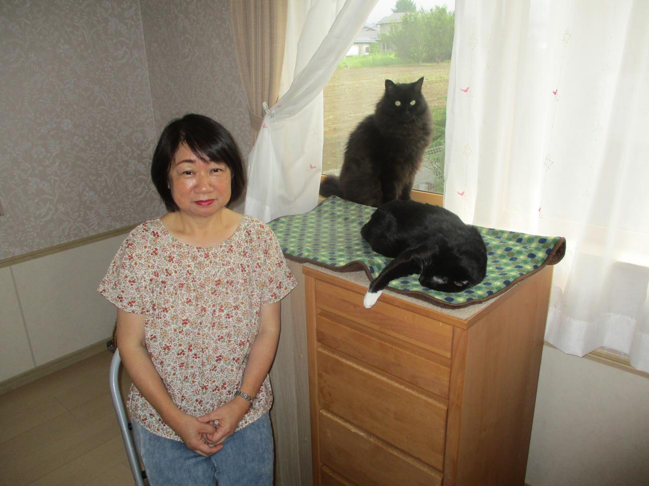 窓の前に座る中川さんと、横の棚にくつろいでいる黒猫2匹の写真