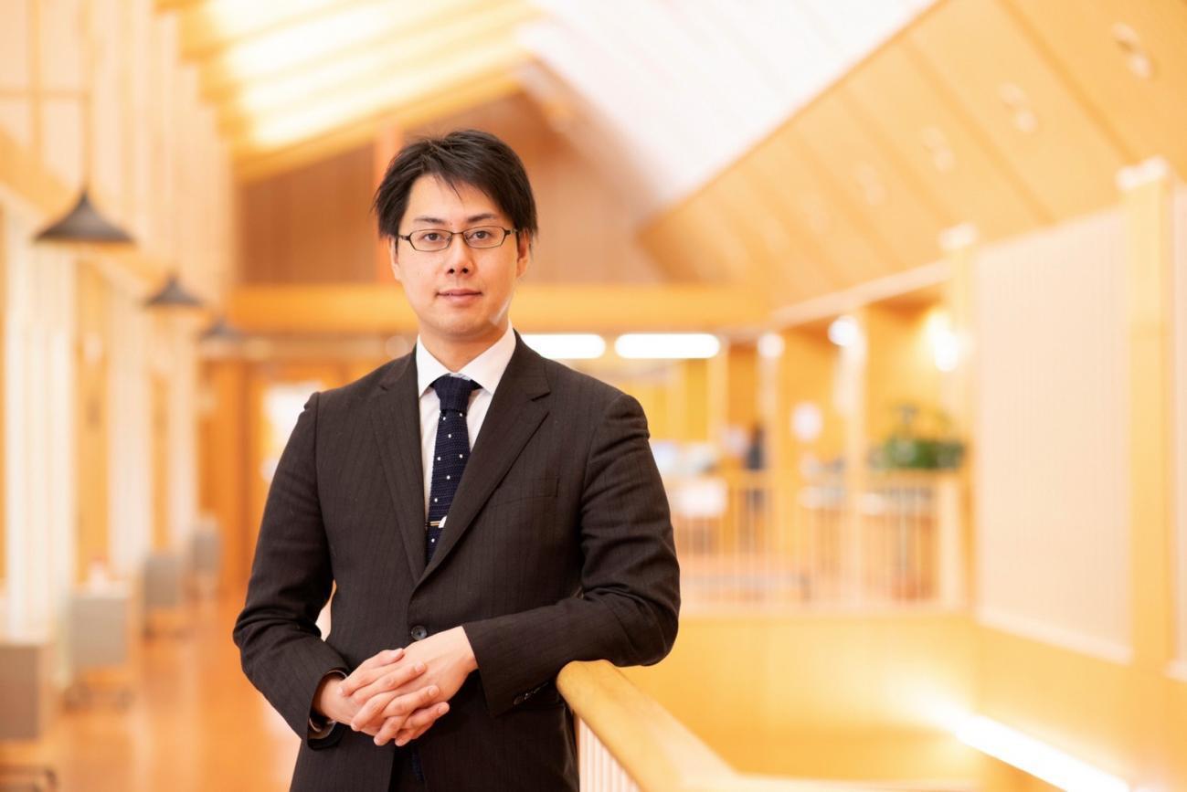 スーツ姿の長谷川 竜也さんが廊下の手すりに肘を置いて、カメラ目線で正面を向いている写真