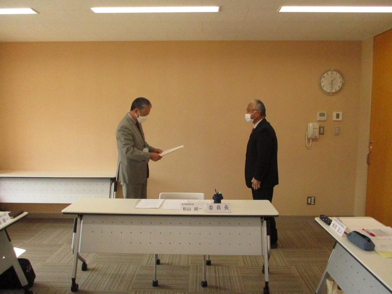 スーツ姿の男性2人が向かい合っており、向かって左側の男性が委嘱状を手に持って読み上げている様子の写真