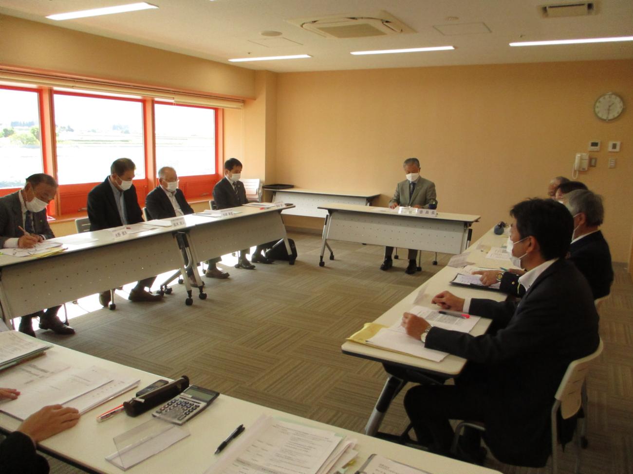 多くのスーツ姿の男性がロの字型に設置された長机に着席し会議をしている写真