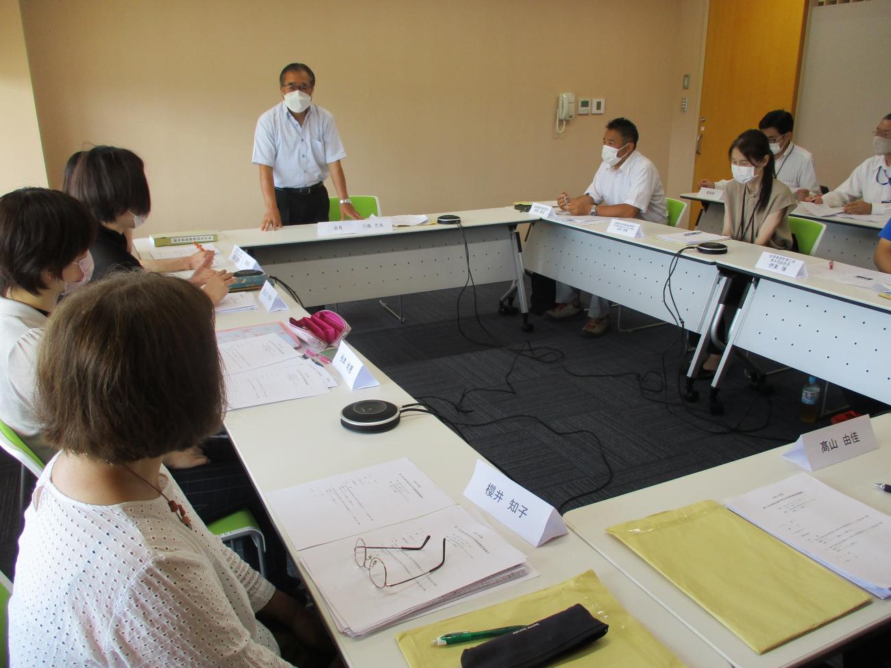 会議室に設置された長テーブルに座って、正面に立っている男性の話を聞いている関係者たちの写真
