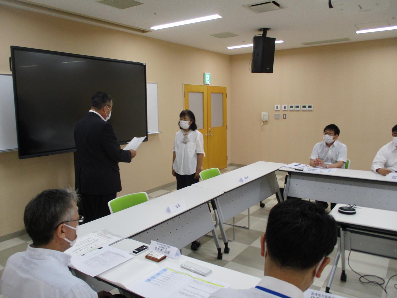 会議室のモニター前で紙を見ながら立っているスーツ姿の男性と、男性と向かい合って立っている女性の写真