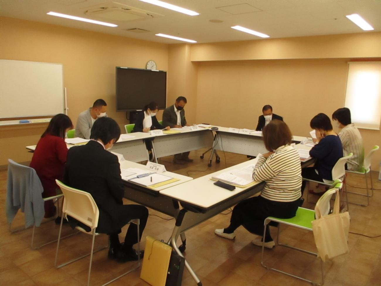 長テーブルがロの字型に設置された会議室で、9人の関係者が手元の資料に目を通しながら会議をしている写真