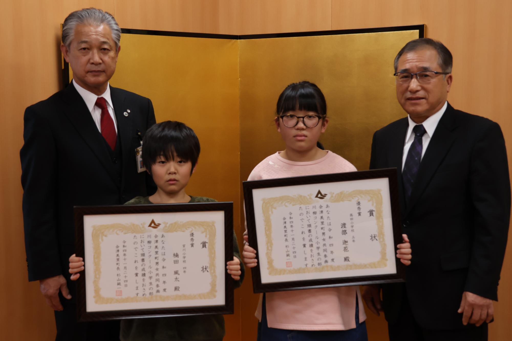 金屏風の前で男子生徒1人と女子生徒1人がそれぞれ賞状を胸の前に持って立っており、町長と川島忠夫会長に挟まれ記念撮影をしている写真