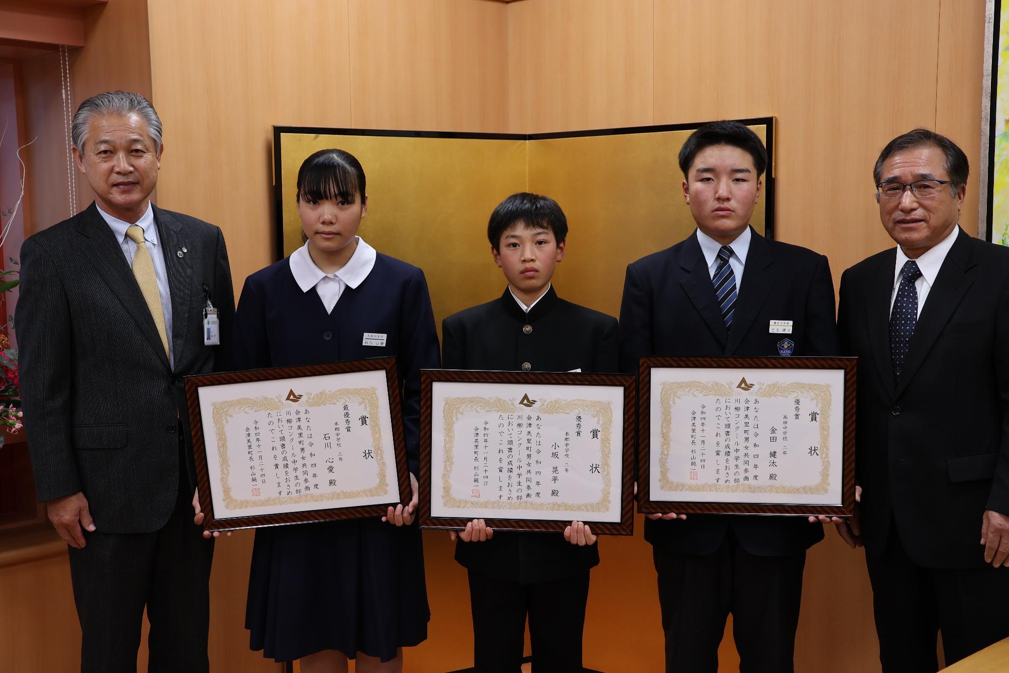 金屏風の前で男子生徒2人と女子生徒1人がそれぞれ賞状を胸の前に持って立っており、町長と川島忠夫会長に挟まれ記念撮影をしている写真