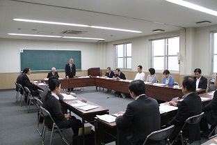 ロの形に設置された長机に多くの関係者が座り、前方の黒板前に立っている男性の話を聞いている様子を会議室の後方から写した写真