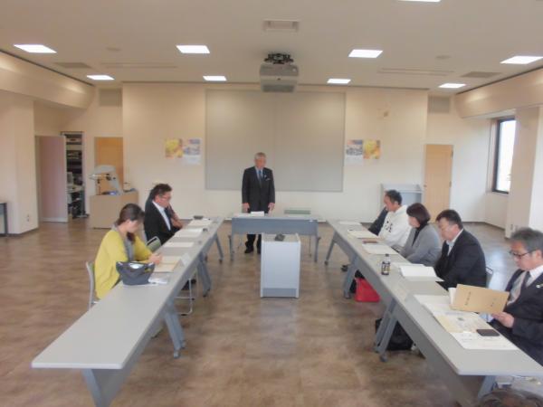 前方にスーツを着た男性が立って話しているのを、コの字型に設置されたテーブルと椅子に座り話を聞いている協議会の様子の写真