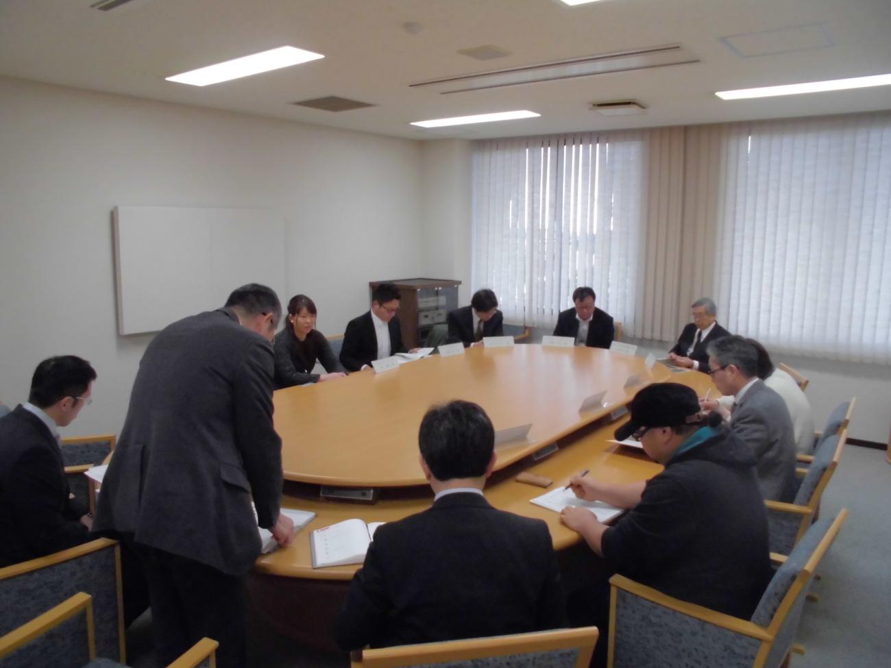 手前にいる男性が立って話をしているのを、楕円のテーブルと椅子に座りメモを取っている協議会の様子の写真