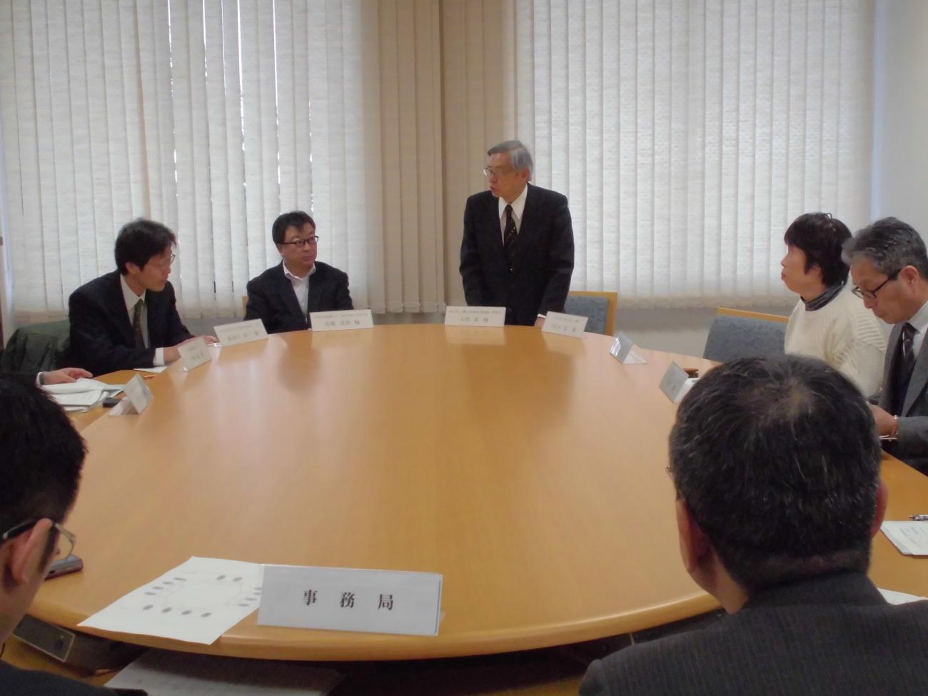 奥にいる男性が立って話しているのを、楕円のテーブルと椅子に座り話を聞いている協議会の様子の写真