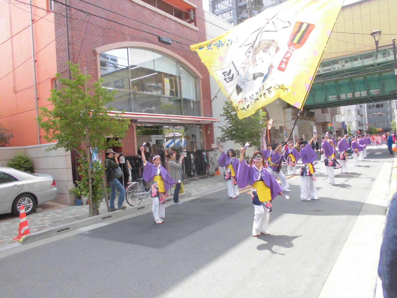 一部の道路上で、紫を基調とした祭り衣装を着た方々が列になって踊り、男性が列の間で大きな旗を持ち振っている写真