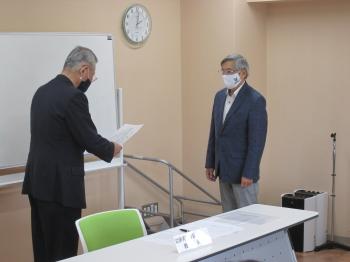紺色のスーツを着た男性（左）が書面を見ながら言い渡しているのを、青色のスーツを着た男性（右）が聞いている写真