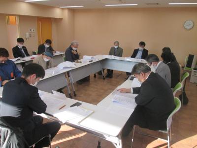複数の男女が、前方に時計が設置された会議室でロの字型に設置されたテーブルと椅子に座り、資料を見ながら話をしている協議会の様子の写真