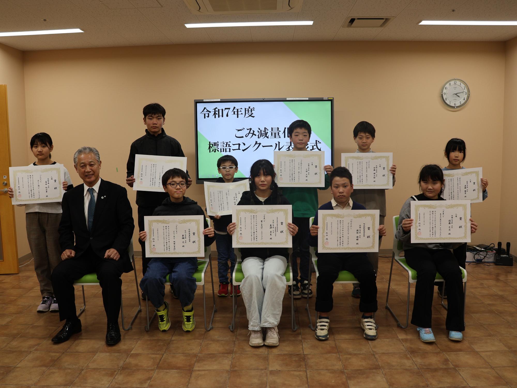 令和7年度ごみ標語コンクールの表彰式で、椅子に座り賞状を見せている9名の受賞者と1名のスーツ姿の男性の写真