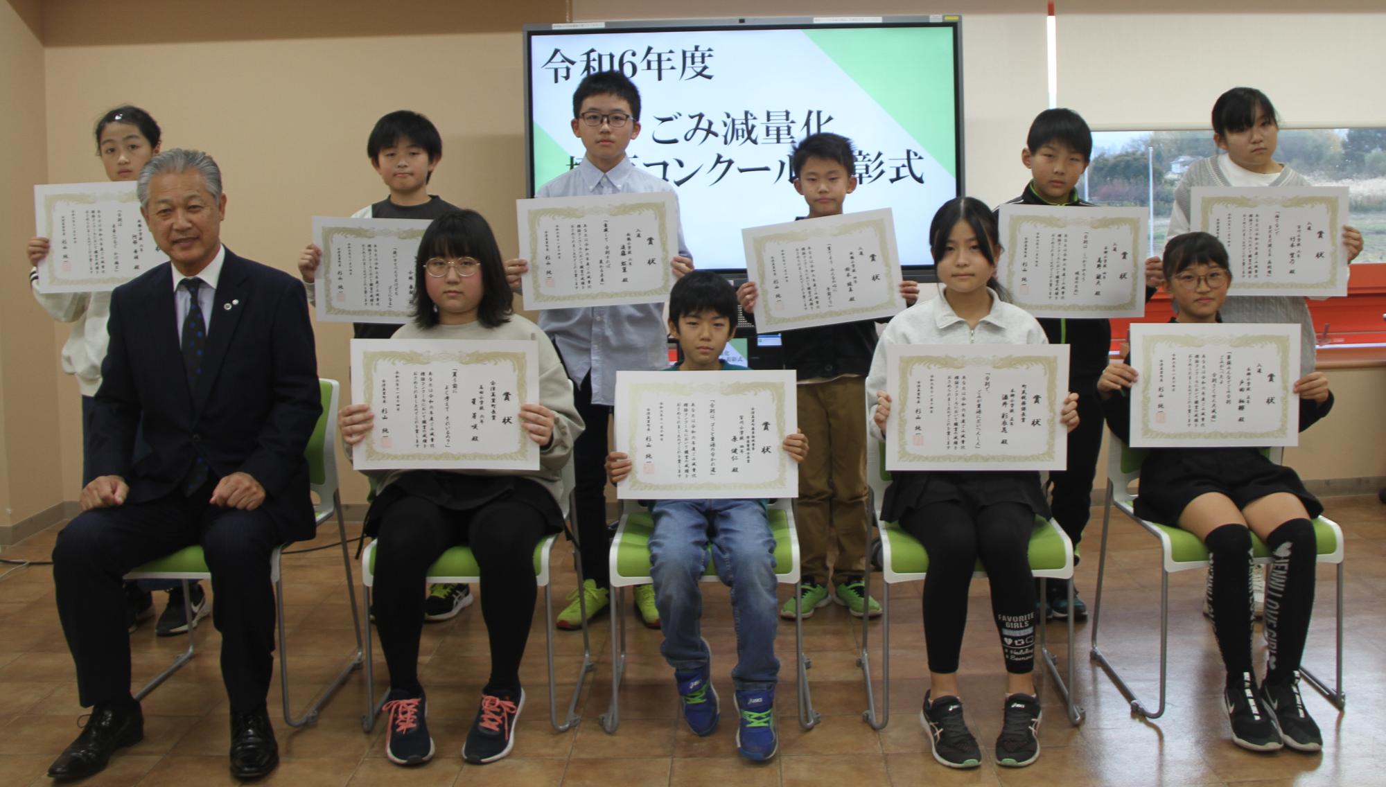 令和6年度ごみ標語コンクールの表彰式で、椅子に座り賞状を見せている9名の受賞者と1名のスーツ姿の男性の写真