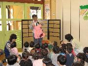 ピンク色のエプロンをつけた女性が前方に立ち、大勢の子供たちに話をしている様子を教室の後ろから写した写真