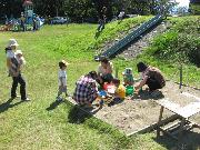 青空の下、公園内の一角にある砂場に小さな子供達と職員の方々が一緒に遊んでいる様子の写真