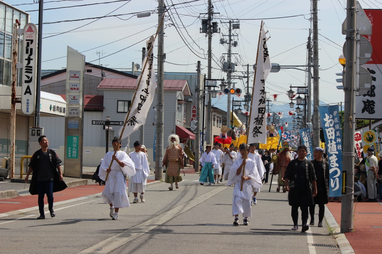 白い祭り衣装を着た方々が列になり、先頭は縦長の旗を持ち、町を練り歩いている写真